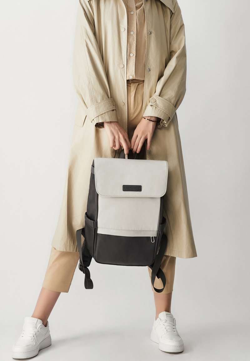 Person i beige jakke og bukser, der holder en sort og lysegrå rygsæk i tophåndtaget, iført hvide sneakers, stående mod en ensfarvet baggrund.