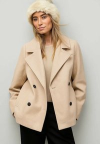 Béžový kabát s dvojitým zapínáním a velkými černými knoflíky, s klopami a strukturovaným designem, doplněný o klobouk z umělé kožešiny.