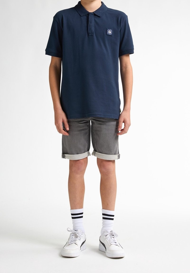 Garçon portant un polo bleu marine, un short gris retroussé, des chaussettes blanches avec des rayures noires et des baskets blanches sur un fond blanc uni.