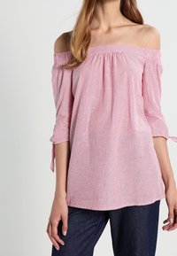 Donna che indossa una blusa a righe rosa e bianca senza spalle con maniche a 3/4 annodate ai gomiti e jeans blu scuro.