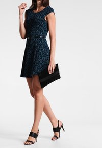 Sukienka z ciemnoniebieskim geometricznym wzorem, krótkie rękawy, podkreślona talia, w połączeniu z czarną kopertówką i odkrytymi czubkami na obcasie. Gładki materiał.