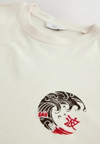 Béžová mikina s krátkym rukávom so zaobleným čiernym a červeným vlnovým vyšívaním a postavou na prednej strane, s jednoduchou textúrou.