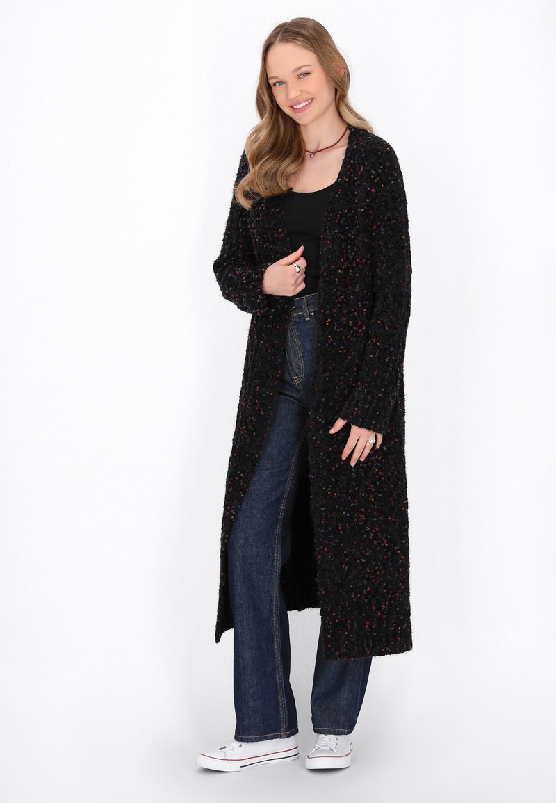 Cardigan lungo nero con specks multicolori, fronte aperto e maniche lunghe, abbinato a jeans blu scuro e sneakers bianche.