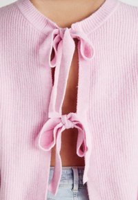 Jasnoróżowy sweter z dzianiny z dwoma związanymi kokardkami z przodu, odsłaniający skórę i jasnoniebieskie dżinsy pod spodem.