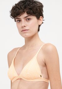 Trójkątny biustonosz bikini w miękkim pomarańczowym kolorze, z teksturowanymi wzorami i cienkimi, regulowanymi ramiączkami. Subtelny złoty akcent na wyposażeniu.
