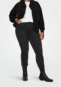 Giacca bomber nera sopra una t-shirt bianca, abbinata a jeans skinny scuri e stivali neri al ginocchio. Focus su una vestibilità rilassata e stratificazione.