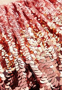 Rosa-gold glänzender Stoff mit runden Pailletten, die wellenförmig angeordnet sind, und einer schimmernden Textur vor einem weichen Mesh-Hintergrund.
