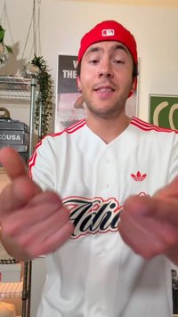 Egy fiatal férfi piros baseballsapkában és fehér Adidas pólóban, aki a kezét a kamera felé nyújtja egy növényekkel és poszterekkel teli szobában.