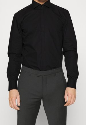Chemise noire à manches longues en tissu lisse, dotée d'un col classique et d'une fermeture à boutons, associée à un pantalon gris sur mesure.