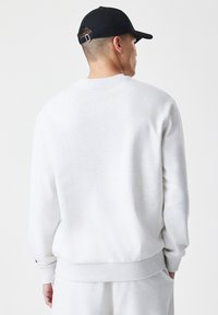 Lys grå sweatshirt med ribbede manchetter og kant, design med rund hals og glat tekstur; båret med en sort cap, set fra ryggen.