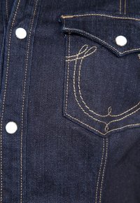 Section de veste en jean bleu foncé montrant une poche avant avec une couture décorative beige et des boutons pression argentés le long de la patte de boutonnage et du rabat de poche.