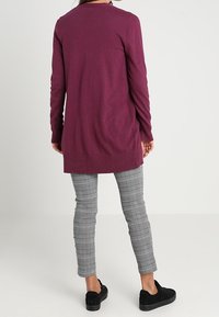 Långärmad, djupt lila kofta i mjukt stickat material, med rak skärning och ribbad nederkant, kombinerad med grå rutig byxor och svarta sneakers.