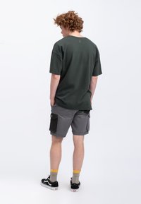 Donkergroen oversized t-shirt en grijze cargoshorts met zijzakken. Shorts hebben een zwart stoffen label en contrasterende stiksels. Zwarte sneakers.
