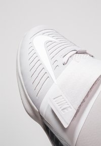 Hvid sportsko med tekstureret overflade, snørebåndsdesign og mærke-stræk. Bemærkelsesværdige detaljer inkluderer perforeringer og et forstærket hælområde.