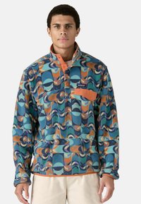 Sudadera de forro polar con patrón geométrico en azul, verde azulado, naranja y crema; presenta un cuello con cremallera de un cuarto y un bolsillo en el pecho con cierre a presión.