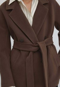 Brązowy, dwurzędowy płaszcz wełniany z paskiem w talii, z dużymi klapami, dwoma guzikami i przeszyciami wzdłuż krawędzi.