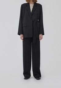 Sort blazer med lange ærmer og bånd i taljen, kombineret med løse sorte bukser. Glat stof, ingen synlige mønstre, klassisk reversdesign.