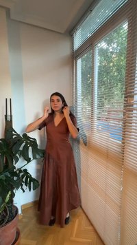 Γυναίκα με καφέ φόρεμα στέκεται δίπλα σε μεγάλο παράθυρο με περσίδες, διορθώνοντας τα μαλλιά της κοντά σε μια γλάστρα με πράσινο φυτό σε ξύλινο πάτωμα.
