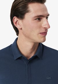 Navyblå polo-shirt gjord av strukturerad tyg med kragn design och en subtil broderad logotyp på bröstet. Tvåknapps-knäppning.