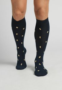 Calcetines negros hasta la rodilla con un patrón repetitivo de pequeñas jarras de cerveza en amarillo y blanco. El material parece suave y elástico.