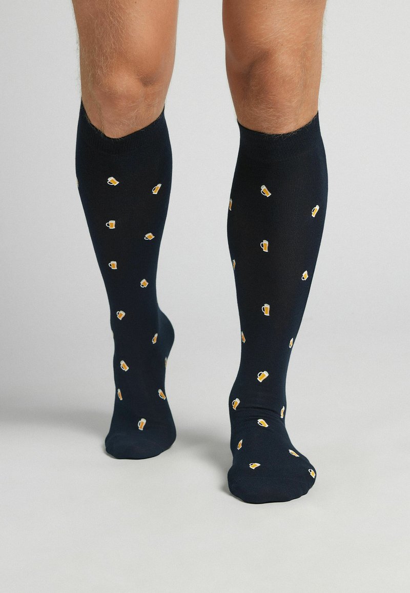 Calcetines negros hasta la rodilla con un patrón repetitivo de pequeñas jarras de cerveza en amarillo y blanco. El material parece suave y elástico.