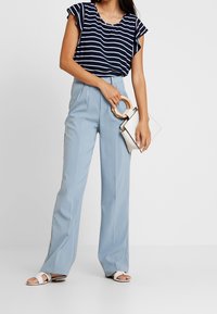 Top a righe blu navy e bianche con maniche corte arruffate, abbinato a pantaloni high-waisted blu chiaro. Borsa bianca con manico di legno.