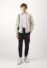 Lys beige knappet skjorte over en hvit t-skjorte, svarte joggebukser med rødt "BULLS"-logo, og hvite sneakers. Svart veske slengt over skulderen.