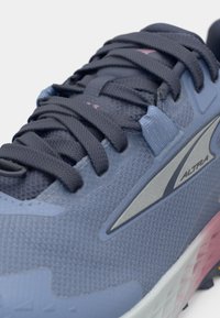 Blue athletic shoe with a textured upper, featuring dark gray laces, a silver accent, and a pink-tinted sole. Rounded toe and cushioned design.