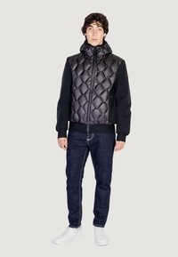 Giacca a vento nera con motivo trapuntato, maniche nere e colletto alto, abbinata a jeans blu scuro e sneaker bianche.
