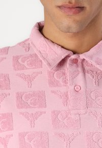 Pink polo shirt featuring raised terry fabric pattern, with square and circular designs, and a buttoned collar.