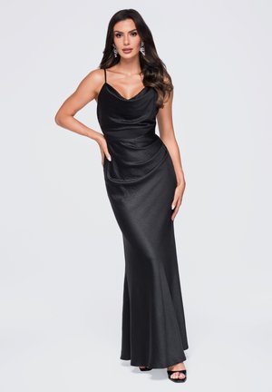 Femme aux longs cheveux foncés portant une robe noire en satin sans manches et des boucles d'oreilles, posant avec une main sur la hanche sur un fond clair.