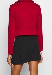 Veste courte rouge à manches longues, confectionnée dans un tissu lisse. Elle présente un large col et une fente au dos, assortie à une jupe noire.