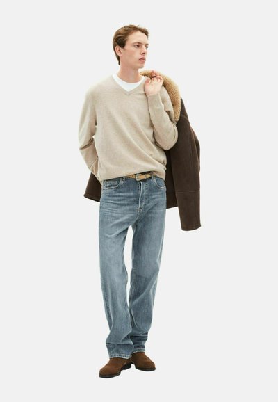 Suéter beige de cuello en v, jeans de mezclilla azules, botas de ante marrón y una chaqueta forrada de piel de oveja marrón colgada sobre el hombro.