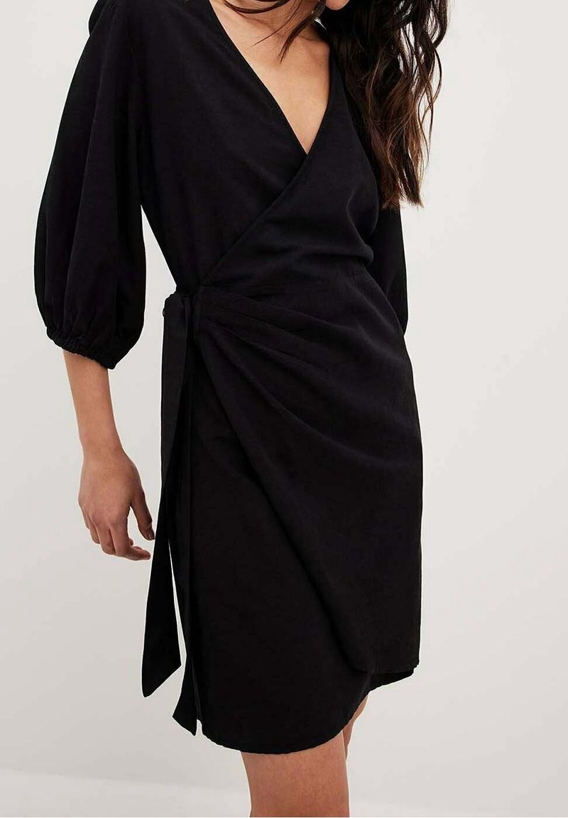 Robe portefeuille noire avec un décolleté en V profond, manches bouffantes et fermeture nouée à la taille, présentant une texture lisse et une coupe jusqu'au genou.