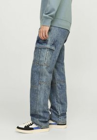 Pantaloni cargo in denim blu medio con diverse tasche applicate, vestibilità ampia, dettagli cuciti e una leggera trama sbiadita. Abbinati a sneaker nere e bianche.