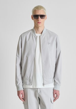 Joven con chaqueta bomber gris claro, camiseta blanca, pantalones cargo gris claro y gafas de sol oscuras, posando frente a un fondo liso.