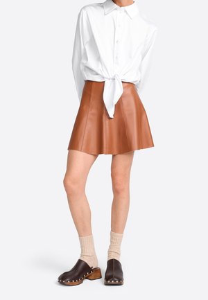 Jupe en cuir marron en forme de A avec plis, associée à une chemise blanche à boutons nouée à la taille, des sabots marron et des chaussettes beige clair.