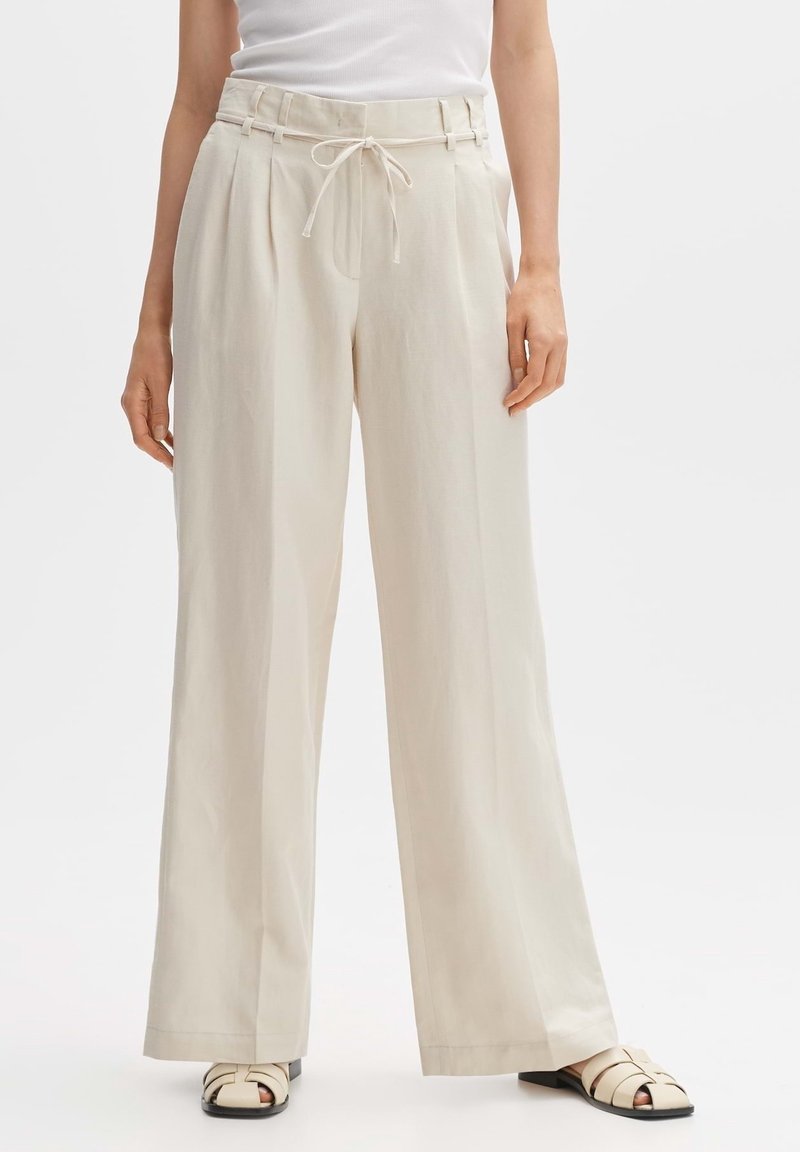 Femme portant un pantalon beige taille haute à jambes larges avec une ceinture à cordon et des sandales blanches, debout sur un fond uni.