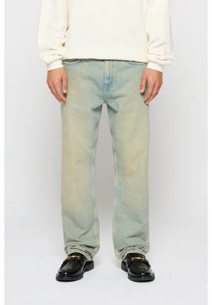 Homme portant un jean bleu clair délavé, un pull côtelé crème, et des mocassins en cuir noir avec un détail de chaîne dorée, debout les mains détendues.