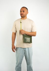 Borsa a tracolla verde oliva con tasca frontale trasparente e tracolla regolabile gialla, indossata da una persona con una maglietta beige e pantaloni azzurri chiari.