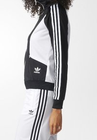 Černobílá sportovní bunda s vysokým límcem, třemi bílými pruhy na rukávech a kapsou na zip. Logo Adidas na hrudi. Měkká, sportovní látka.