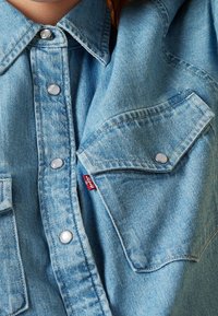 Vaaleansininen farkkupaita, jossa on terävä kaulus, painonapit ja rintatasku, jossa on pieni punainen etiketti. Pinta on sileä, ja siinä on haalistuneita kohtia.