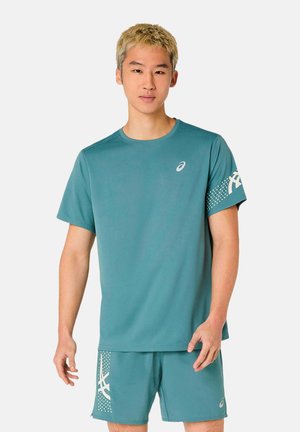 Giovane uomo con capelli biondi corti che indossa abbigliamento sportivo Asics turchese, composto da una maglietta a maniche corte e pantaloncini abbinati con dettagli del logo bianchi.
