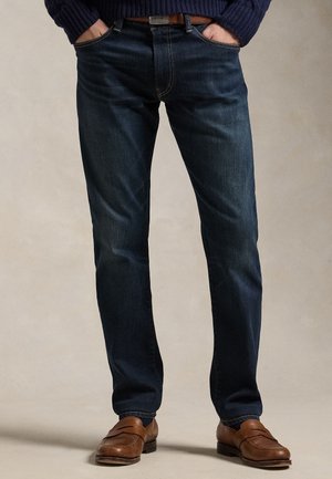 Άνδρας που φορά σκούρο μπλε τζιν και καφέ δερμάτινα λουφάρια, με τα χέρια στις τσέπες, στέκεται μπροστά σε ουδέτερο φόντο.