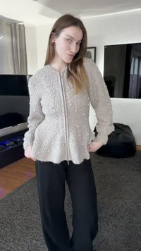 Grijze gebreide cardigan met een rits aan de voorkant, gestructureerd ribbelpatroon en verspreide witte pomponaccenten. Gecombineerd met zwarte broek met wijde pijpen.