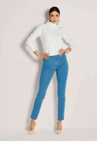 Witte kabelgebreide turtleneck trui gecombineerd met lichtblauwe skinny jeans. Model draagt beige puntige hakken. Heldere achtergrond.