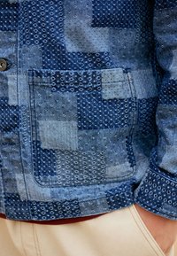 Tasca e manica di giacca di jeans patchwork con cuciture strutturate, indossata sopra pantaloni beige con mano nella tasca.