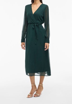 Kobieta ubrana w ciemnozieloną sukienkę midi zapinaną na zakładkę z przezroczystymi długimi rękawami oraz złote sandały na wysokim obcasie z paskami, stojąca na białym tle.