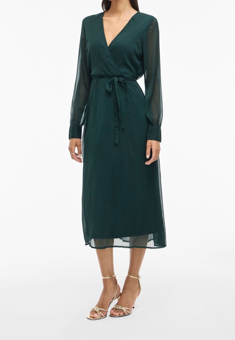 Femme portant une robe portefeuille midi vert foncé avec manches longues transparentes et sandales à talons hauts dorées à brides, debout sur un fond blanc.