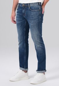 Jean en denim brut avec une coupe slim, doté d'une fermeture à bouton, de poches avant et arrière, et de revers. Associé à des baskets blanches.